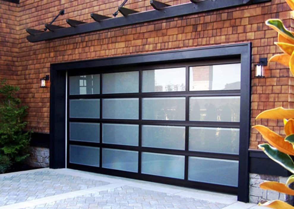 Exterior view of a modern full-view glass garage door with a black aluminum grid frame, integrated into a house facade with warm cedar shingle siding and a stone driveway.