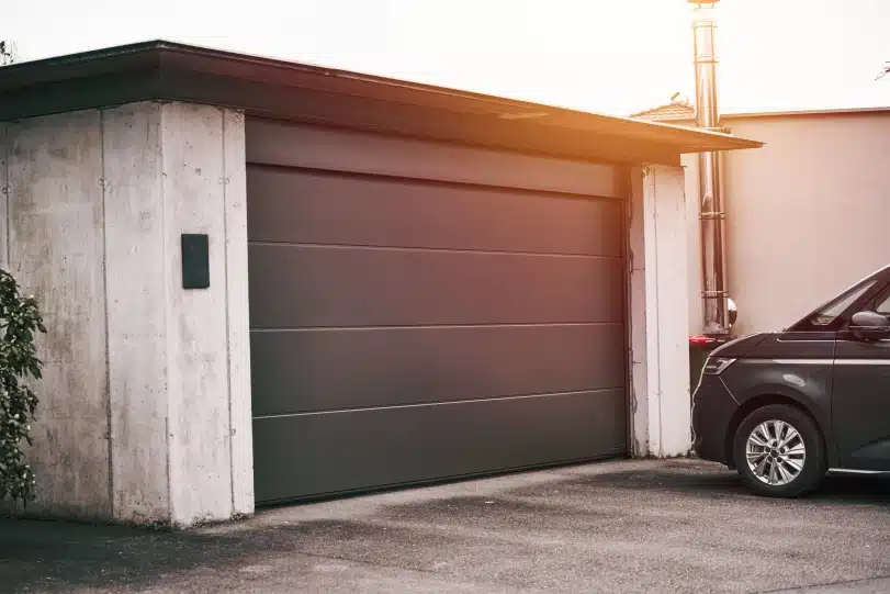 Modern charcoal gray flush panel sectional garage door installed on a concrete residential garage structure with a dark van parked in the driveway.