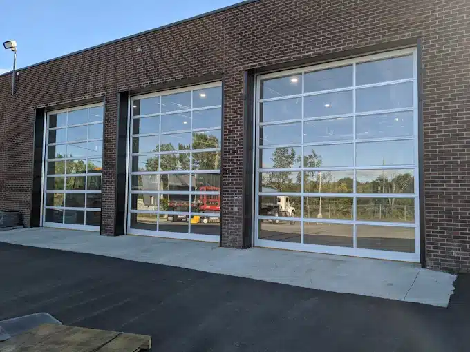 Three large full-view commercial glass garage doors with white aluminum frames installed in a modern dark brick building bay.
