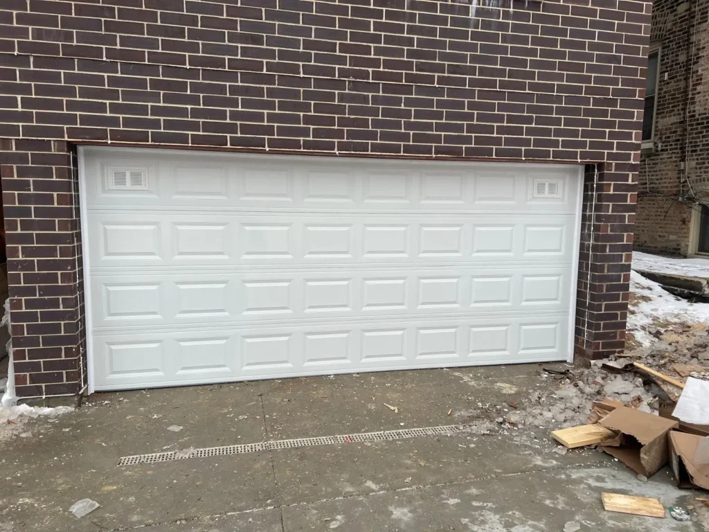 A wide, white double-car garage door with a raised panel design and corner ventilation inserts installed in a dark brick facade.