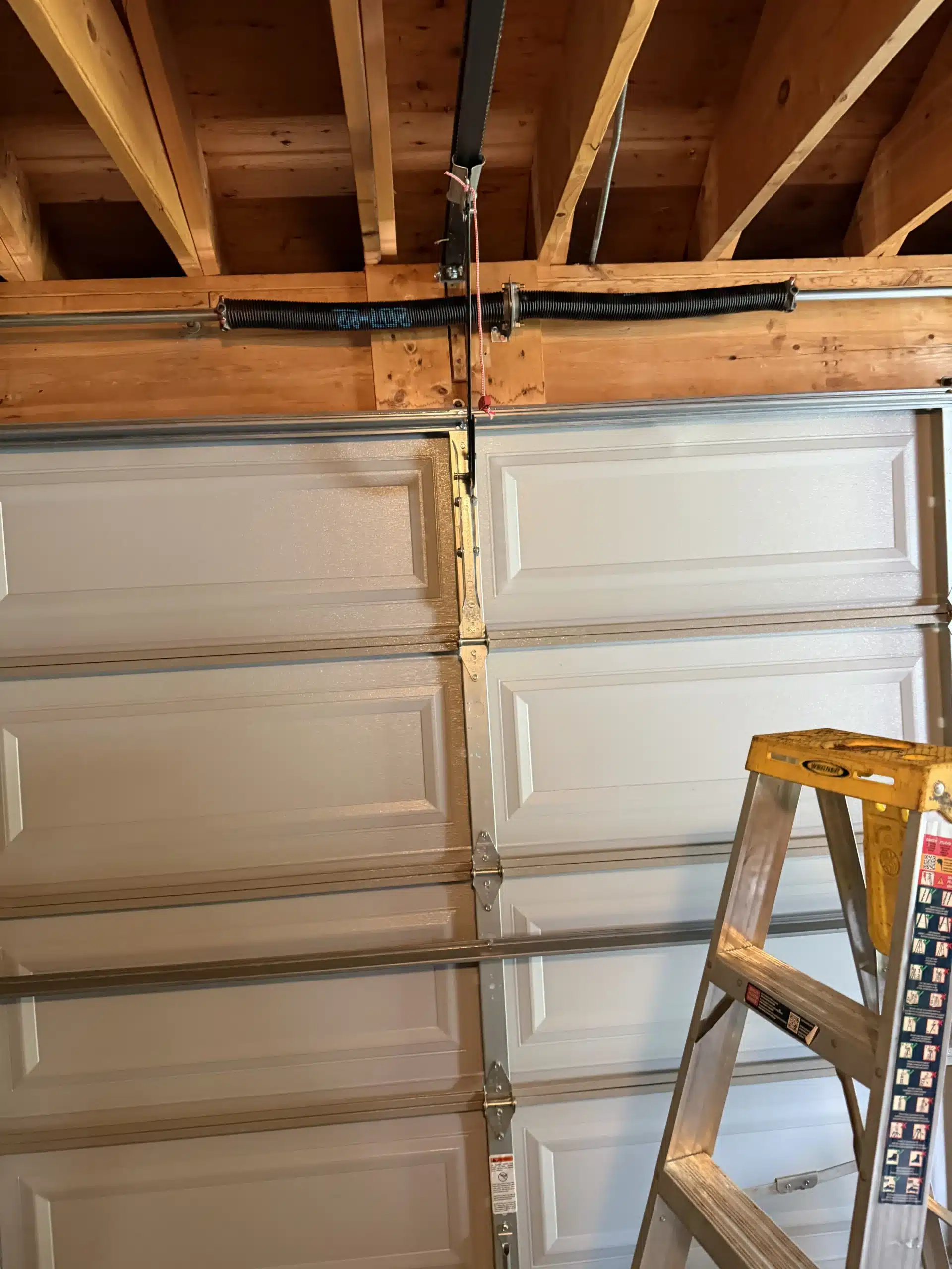 Interior view of a mounted white paneled garage door showing overhead torsion springs, vertical tracks, and a step ladder.