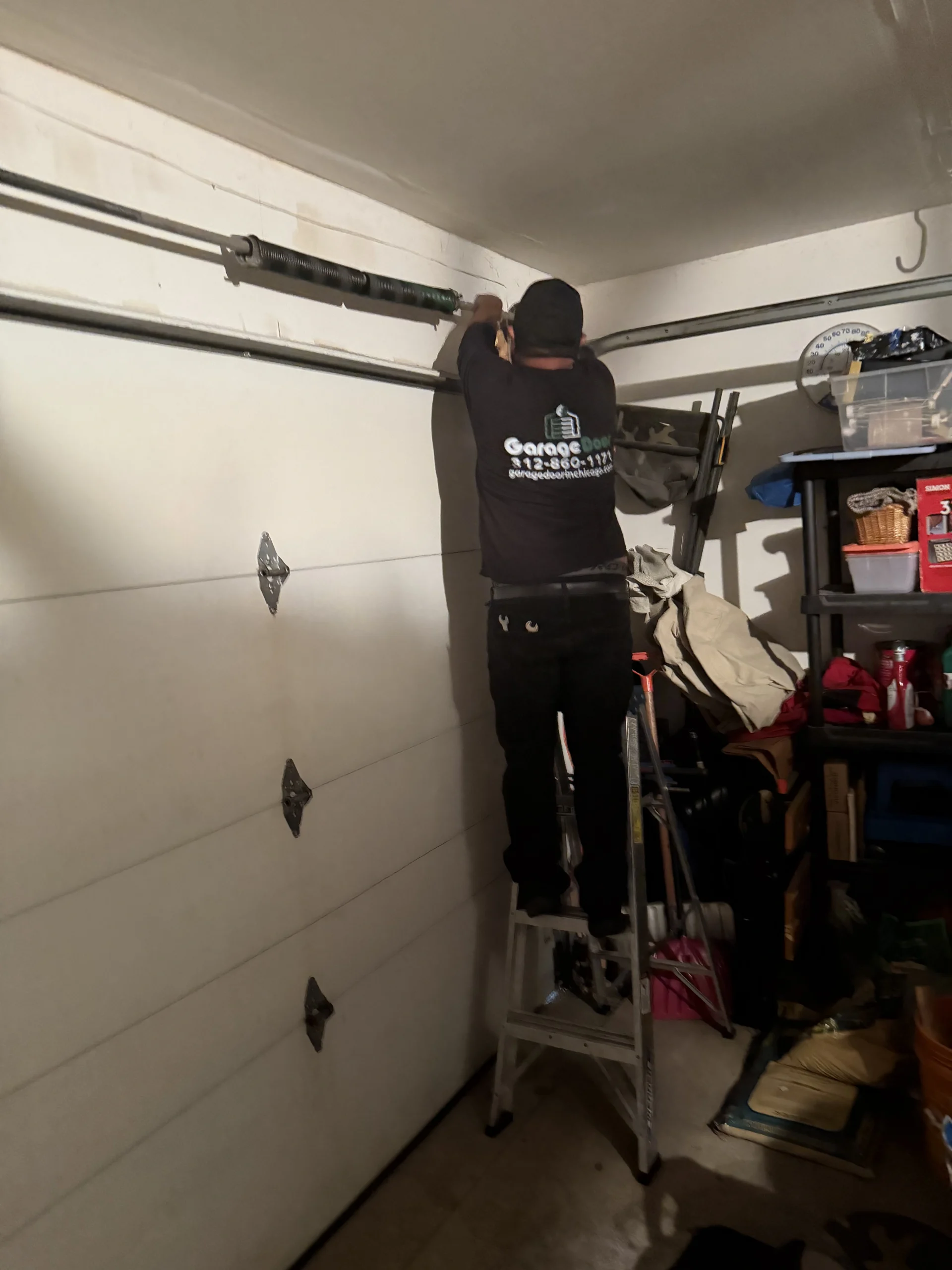 A technician standing on a ladder and adjusting the upper spring mechanism of a closed garage door.